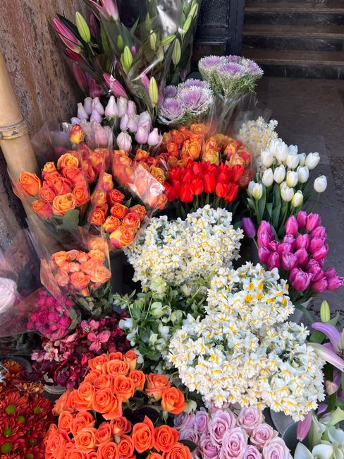 A vibrant assortment of fresh flowers arranged in bouquets and clusters, featuring orange, pink, white, and purple blooms. The display includes roses, tulips, and hydrangeas, with some wrapped in clea