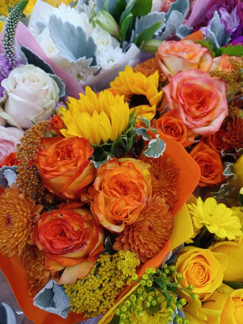 A vibrant floral arrangement featuring orange roses, yellow chrysanthemums, and softened pink roses, complemented by sprigs of green foliage and greyish-green dusty miller leaves. The bouquet is wrapp