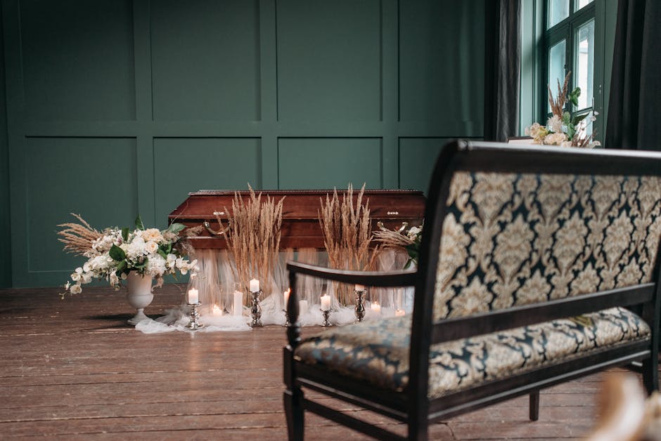 A funeral setting featuring a closed wooden casket adorned with surrounding floral arrangements. To the left, a white ceramic vase holds a bouquet of white roses, lilies, and greenery, contrasted by t