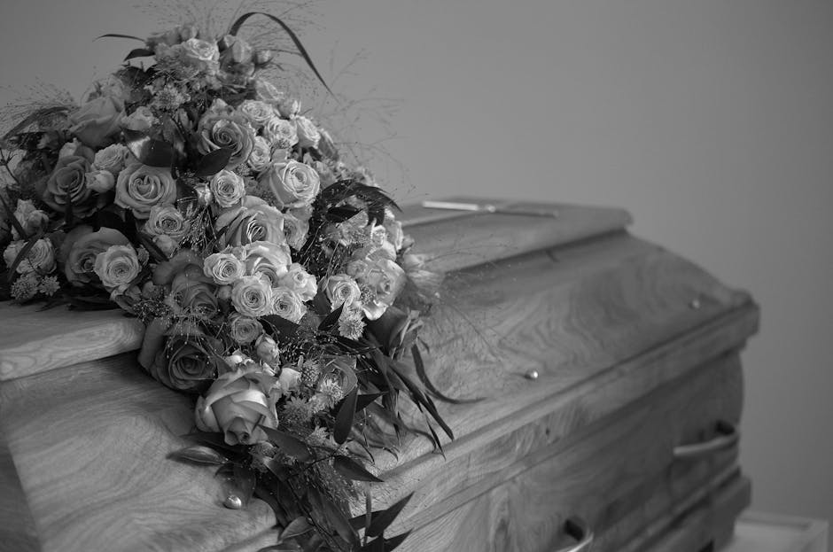 A bouquet of various fresh flowers placed on top of a closed wooden casket. The arrangement includes roses and smaller blossoms, with some greenery interspersed. The flowers are in shades of light and