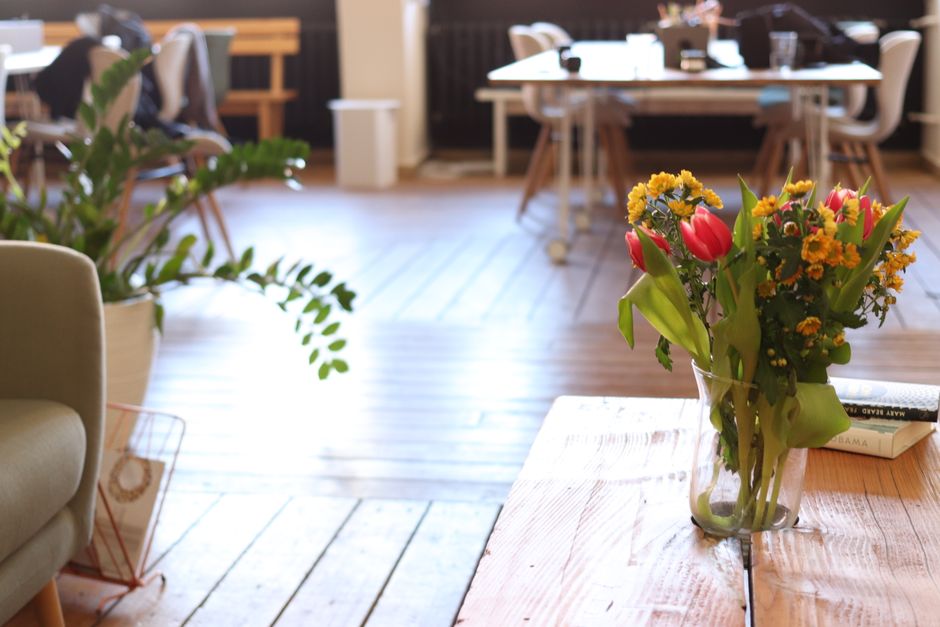 A clear glass vase containing a floral arrangement of red tulips, small yellow and orange daisies, and green foliage sits on a wooden table inside a well-lit, modern interior space. The bouquet appear