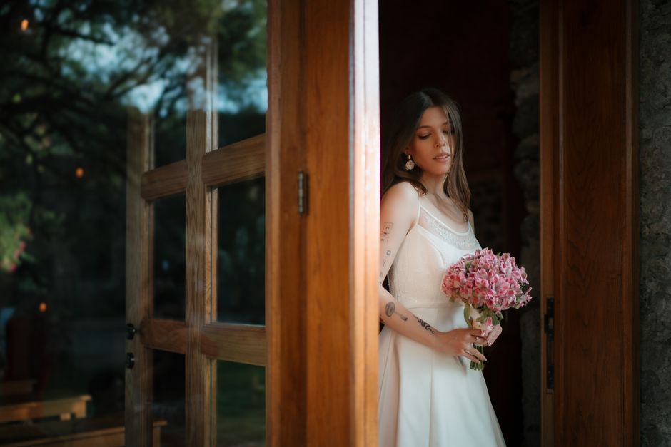 A young woman with shoulder-length brown hair, wearing a sleeveless white dress with lace detail, stands partially behind a wooden doorframe holding a bouquet of pink hydrangeas. The bouquet is fresh,