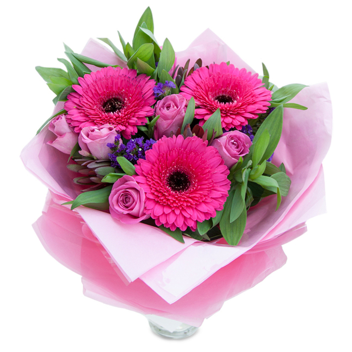 I see this arrangement as a balance of confident colour and gentle romance, centred on three vivid fuchsia Gerbera daisies whose many petals create a striking, almost graphic presence against the softer blooms. Surrounding them are roses in a dusty lavender-pink, their tight, velvety petals suggesting tenderness and a lingering, discreet perfume that does not overwhelm. Small sprays of purple statice float between the larger flowers, adding pockets of texture and a cool tonal contrast that gives the eye places to rest. The foliage is generous and green-leaves with the glossy heft of salal and the subtle scent of eucalyptus weave through the stems, creating a lush, garden-like foundation that amplifies the intensity of the pinks. The bouquet is wrapped in layers that read as thoughtfully chosen: a matte pale-pink tissue lining cradles the stems and softens the presentation, while an outer sheet of translucent fuchsia-pink cellophane, slightly crinkled, lends a delicate shine and cohesion to the pink palette. The clear glass vase rim peeking from the lower wrap signals readiness for display and the freshness of the stems. Imagining this on a small dining table in a Castelnau flat or set by the window of a cottage near Barnes Bridge, the arrangement feels like a warm, considered gift: suitable for birthdays, anniversaries, or an impulsive expression of affection. The overall mood is buoyant yet intimate, the kind of bouquet a local florist would wire with care and deliver to bring a moment of colour and calm to a neighbour.