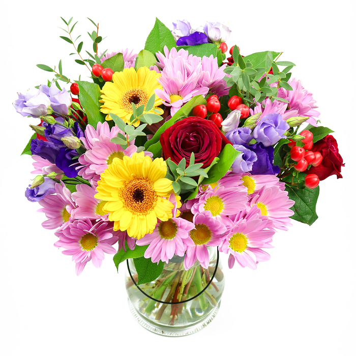 A clear glass vase sits centrally in the frame, brimming with an abundant bouquet that feels both polished and delightfully natural, as if gathered from a well-tended garden just off the leafy avenues of Castelnau. In the centre, velvety red roses spiral open, their deep, romantic hue anchoring the design and giving it a sense of classic sentiment. Flanking them are two vivid yellow gerbera daisies, their broad petals stretching out from dark, warm centres like rays of sunshine, instantly adding a bright and friendly character. Around these statement blooms, soft pink daisies or chrysanthemums form generous clusters, each flower with many slender petals and a neat yellow centre, creating a halo of pastel colour that gently softens the stronger tones. Rising among them, elegant lisianthus in purple and pale lavender bring a cool contrast; their delicate, bell-shaped blooms and graceful stems lend a modern, almost airy sophistication. Tucked in between petals, you can spot round red hypericum berries, glossy and playful, adding an extra layer of texture and a touch of whimsy. The whole arrangement is woven through with lush green foliage-some leaves broad and deep green, others finer and lighter-giving the bouquet structure while keeping it relaxed and full of movement. Through the transparent vase, carefully trimmed stems are visible, arranged neatly in clear water, a small yet telling detail of the florist's craftsmanship. The atmosphere this arrangement creates is joyful and welcoming, ideal as a cheerful centrepiece for a family table near Barnes Village, a thoughtful birthday surprise, or a colourful congratulations bouquet delivered locally by Castelnau Florist to friends and neighbours along the river.